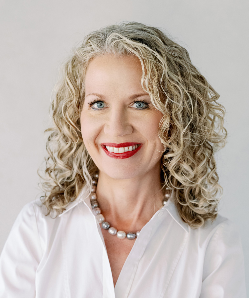 Smiling woman with curly blonde hair and pearl necklace in white blouse.